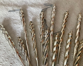 Könnte beinhalten: Eine Sammlung von silberfarbenen Vintage-Weihnachts-Eiszapfen-Ornamenten. Die Ornamente haben ein gedrehtes, seilartiges Design und werden von kleinen Metallhaken gehalten. Die Eiszapfen sind auf einer weißen, strukturierten Oberfläche angeordnet.