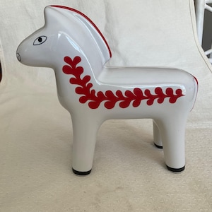 May include: A white ceramic horse figurine with red detailing. The horse features a red mane and a red floral pattern along its side. The figurine has black hooves and a simple, stylized design.
