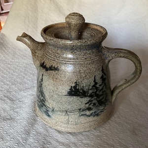 May include: A speckled ceramic teapot with a lid and handle. The teapot features a winter landscape scene in blue and black. The spout is on the left, and the handle is on the right. The teapot is a rustic design.