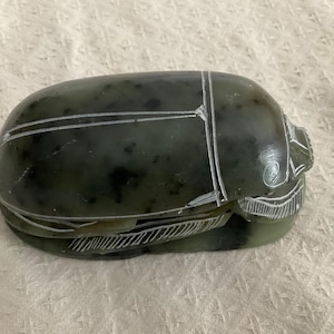 May include: A dark green scarab beetle figurine with white etched details. The beetle is oval-shaped with carved lines and markings, and is likely made of stone or a similar material. The figurine has a smooth, polished surface.