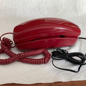 May include: A glossy dark red landline telephone with a matching coiled cord and a separate black straight power cord. The handset rests horizontally on the base unit, featuring a smooth, curved plastic exterior and a simple, functional design.