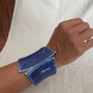 May include: A wide cuff bracelet featuring two curved, rectangular panels with a glossy, deep blue enamel finish and visible brushstroke textures. The metal edges are silver-toned, framing the blue surfaces which meet at a central open gap.