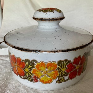 Peut inclure: Plat à gratin en céramique blanche avec un couvercle. Le plat est décoré d'un motif floral dans les tons orange, jaune et vert. Le couvercle a un bouton sur le dessus.