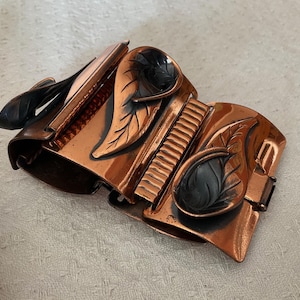 May include: A vintage copper bracelet with a leaf design. The bracelet features multiple hinged sections with detailed leaf and stem accents. The leaves are a dark, contrasting colour against the copper. The bracelet is a statement piece.