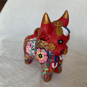May include: A hand-painted ceramic bull figurine. The bull is mainly red with gold-tipped horns and accents. Floral patterns in pink, green, and white decorate the body. The figurine has a whimsical, folk art style.