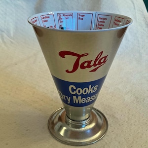 Peut inclure: Un ancien verre doseur sec Tala. La tasse argentée a une forme de cône évasé avec une bande bleue et un texte rouge qui dit "Tala Cooks Dry Measure". Le bord comporte des mesures imprimées pour divers ingrédients. La base est un petit piédestal rond argenté.