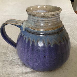 May include: A round ceramic mug with a wide base and a narrow neck. The lower portion is a deep purple, transitioning into a light blue drip pattern. The upper rim and handle are a neutral beige colour with a smooth, glossy finish throughout.