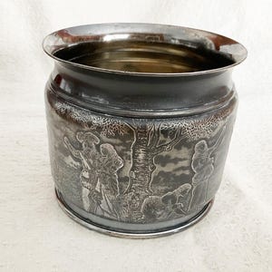 Peut inclure: Un récipient en métal argenté avec une scène gravée de personnages dans un paysage. Le récipient a une large ouverture et un bord et une base polis. La gravure détaillée entoure l'extérieur. Un objet décoratif.