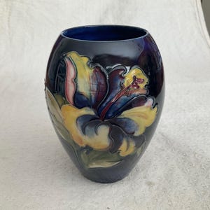 May include: A dark blue ceramic vase with a glossy finish, featuring a raised, embossed floral design in shades of yellow, pink, and deep purple. The vase has an oval, rounded shape with a wide opening and a smooth, reflective surface texture.