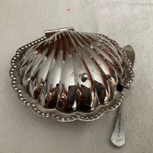 May include: A silver-toned metal serving dish shaped like a scallop shell with a hinged lid and a beaded edge. A small matching silver-toned metal spoon with a flat handle is tucked underneath the shell, resting on the surface beside the dish.
