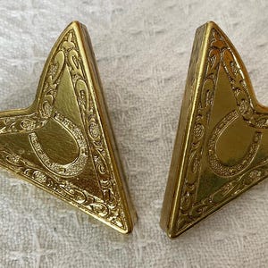 May include: Two gold-coloured, triangular decorative objects. Each has a horseshoe design in the centre and ornate scrollwork around the edges. The objects are displayed on a white, textured surface.