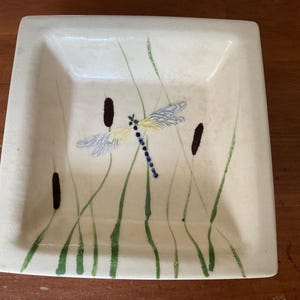 May include: Square ceramic dish with a hand-painted dragonfly and cattails design. The dragonfly has blue, yellow, and white wings with a blue body. Green blades of grass and brown cattails are also painted on the off-white surface.
