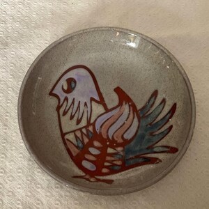 May include: A small, round ceramic bowl featuring a hand-painted bird design. The bird is rendered in red, blue, purple, and orange hues against a speckled grey backdrop. The bowl's rim is a darker shade of grey.