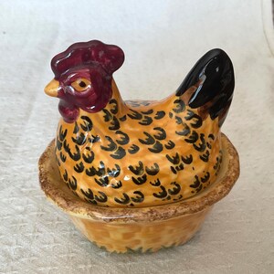 以下が含まれることがあります： お揃いのボウルに入った陶器製の鶏の置物。鶏は黒い羽模様のある黄色い体、濃い赤色のトサカと肉垂、黒い尾を持っています。ボウルは質感のある茶色の斑点模様の縁と、黄色の土台で構成されています。全体的に光沢のある仕上げが施されています。