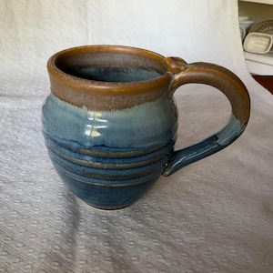 May include: A ceramic mug with a rounded body and a large handle. The mug has a gradient of colours, with a brown rim, a light blue middle, and a dark blue base. Horizontal lines encircle the body.