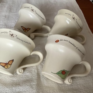 May include: Four cream-coloured ceramic mugs with curved handles. Each mug features a butterfly or ladybug design with the words "beautiful dancing butterflies" or "the most humblest of nature's creatures".