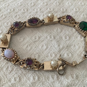 May include: A gold-tone link bracelet featuring alternating rectangular metal segments and decorative settings. The bracelet is adorned with round white pearls, faceted purple stones, a single iridescent opal-like cabochon, and a green stone.