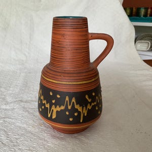 May include: A vintage ceramic vase with a handle. The terracotta-coloured body has horizontal brown stripes. A dark brown band with yellow and gold abstract designs encircles the vase. The interior is teal.