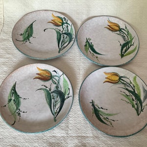 Peut inclure: Quatre assiettes rondes en céramique avec une surface blanc cassé mouchetée. Chaque assiette présente un motif peint à la main d'une tulipe jaune avec des feuilles vertes et un bord bleu clair. Les assiettes sont disposées sur une surface blanche.