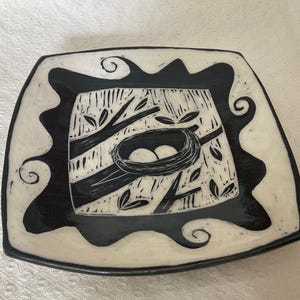 May include: Square ceramic plate with a black and white design. The plate features a central image of a bird's nest with two eggs, surrounded by branches and leaves. The border is black with a wavy, decorative pattern.