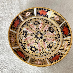 May include: A small, round, decorative plate with a blue and gold floral pattern. The plate has a red and gold border.