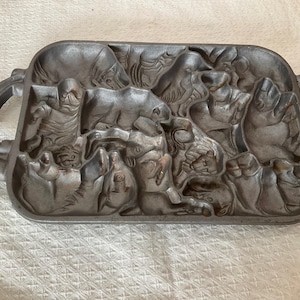Peut inclure: Un moule à pâtisserie rectangulaire en fonte avec un motif en relief détaillé de divers animaux et personnages. Le moule a deux poignées arrondies et une finition gris foncé. Le motif comprend des ours, des oiseaux et d'autres créatures.