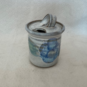 May include: A white ceramic jar with a lid and a blue and green floral design. The jar has a handle on the lid.