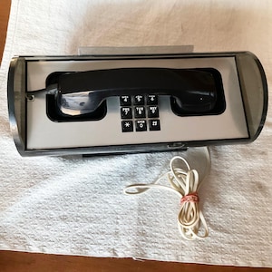 May include: A retro black telephone with a rectangular keypad and a black handset. The phone is housed in a clear, curved plastic casing with a silver faceplate. A coiled white cord is attached.