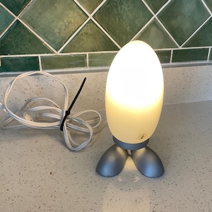 Peut inclure: Une lampe de table dotée d'un abat-jour en verre dépoli blanc en forme d'œuf qui émet une lueur chaude. La base est constituée de deux pieds arrondis de couleur argentée. Un cordon électrique blanc avec un serre-câble noir est fixé à la base.