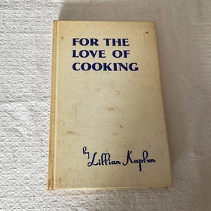 以下が含まれることがあります： リリアン・カプラン著のヴィンテージ料理本「FOR THE LOVE OF COOKING」。本はクリーム色の表紙に青色の文字が書かれています。表紙には使用感があり、愛されている料理本であることがわかります。