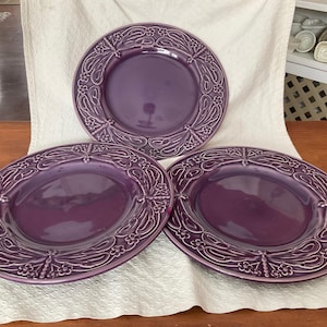 May include: Three oval purple ceramic plates with detailed floral and scrollwork patterns. The plates have a glossy finish. They are arranged on a white, textured cloth on a wooden table. Each plate has a decorative border.