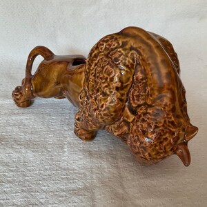 May include: A brown glazed ceramic planter shaped like a bison. The figure features a textured, curly mane and a smooth body, with an opening on the back for plants. The bison is depicted in a lowered head position, showcasing a glossy finish.