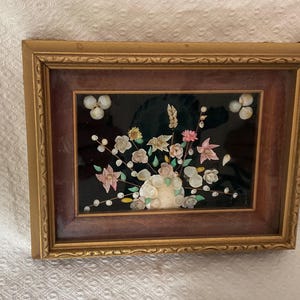Peut inclure: Une décoration murale rectangulaire présentant un arrangement floral composé de divers petits coquillages dans des tons de blanc, rose et vert, sur un fond noir, encadré par un cadre en bois foncé avec une bordure extérieure dorée.