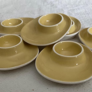 May include: A set of six pale yellow ceramic egg cups, each featuring a wide, rounded base and a central circular holder with a white rim. The cups are arranged in a cluster, showcasing their smooth, glossy finish and consistent design.