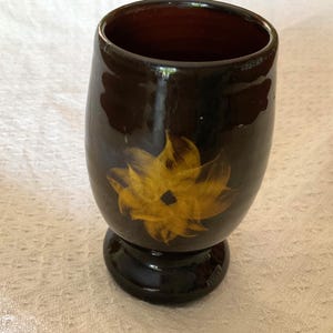 May include: A dark brown ceramic cup with a pedestal base. The cup features a yellow floral design on the front. The interior of the cup is a lighter brown colour. The cup has a glossy finish.