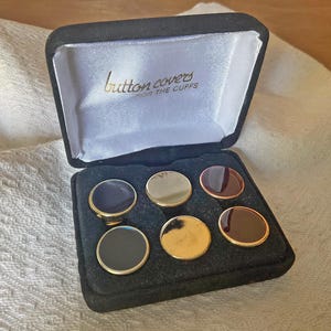 May include: A set of six circular button covers for cuffs housed in a black velvet-lined presentation box. The interior lid features the text button covers FOR THE CUFFS. The covers include various metallic and solid coloured circular designs.