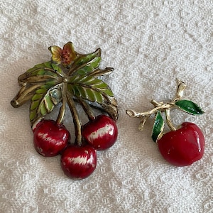 Peut inclure: Deux broches vintage en forme de cerises. L'une représente trois cerises rouges avec des reflets blancs et des feuilles vertes. L'autre a une seule cerise rouge avec des feuilles vertes. Les deux broches ont des accents métalliques dorés.