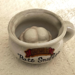 May include: A white ceramic miniature chamber pot with a handle, featuring a sculpted human posterior inside. The exterior displays a red banner with the text SOUVENIR OF CORPUS CHRISTI, TEX and the black printed text Butt Snuffer.