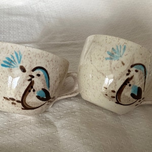 May include: Two white ceramic coffee mugs with brown and blue bird designs. The mugs have a speckled glaze and a handle.