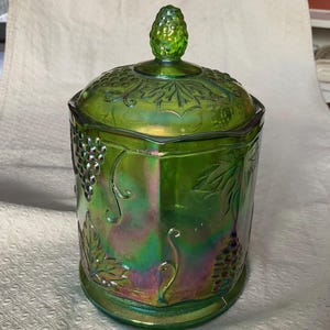 May include: Green iridescent glass jar with a lid. The jar features embossed grape and leaf designs. The lid has a knob and the jar has a hexagonal shape. The glass has a shiny, reflective surface.