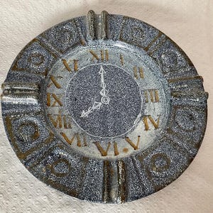 Peut inclure: Cendrier rond avec un motif de cadran d'horloge. Le cendrier présente des chiffres romains et des aiguilles indiquant 8 heures. Le bord extérieur est texturé avec une finition mouchetée bleue et marron.