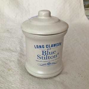 以下が含まれることがあります： 蓋付きの白い陶器の瓶。瓶には「LONG CLAWSON Blue Stilton」と「Stilton makers for over 100 years」と書かれた青い文字があります。瓶は円筒形で、底が丸く、蓋に小さなノブが付いています。