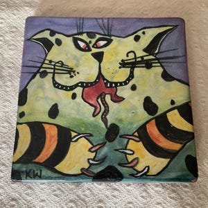 May include: Square ceramic tile with a quirky cat illustration. The cat is depicted with yellow fur, black spots, red eyes, and a red tongue. The tile has a purple and green background, and the artist's initials, "KW", are in the corner.