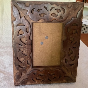 May include: A rectangular wooden picture frame featuring a dark brown finish and intricate, hand-carved openwork patterns along the border. The centre contains a plain, light brown backing board with two small, circular metal fasteners visible.