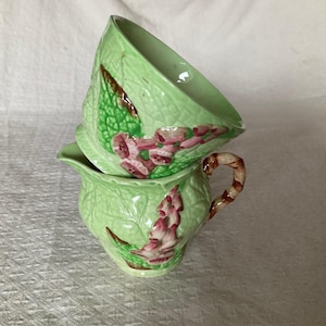 Peut inclure: Ensemble en céramique vert clair comprenant un pot à crème avec anse et un sucrier assorti. Les deux pièces présentent un motif de feuilles avec des accents roses et bruns. Le pot à crème mesure environ 10 cm de haut.