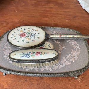 May include: A vintage dressing table set comprising a hand mirror, a brush, and a small brush, each with floral embroidery on a cream surface. The set is displayed on a decorative tray with a lace design and metal edging.