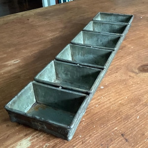 May include: A rectangular metal baking tin featuring five connected individual compartments arranged in a single row. The tin has a weathered, dull grey finish with visible signs of oxidation and wear along the edges and interior surfaces.