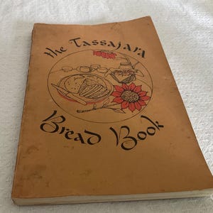 May include: A softcover book titled The Tassajara Bread Book with a tan cover. The front features a circular illustration of sliced bread, a knife, a basket, and a large red flower. The text is printed in a black, stylised, cursive-like font.