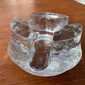 May include: A clear, textured glass candle holder with three curved sections and a central well. The design features a unique, organic shape, perfect for displaying candles or small decorative items. The glass has a slightly frosted appearance.