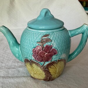 May include: A turquoise ceramic teapot with a lid, spout, and handle. The body features a diamond pattern and a raised floral design with burgundy and yellow accents. The teapot is a decorative kitchenware item.